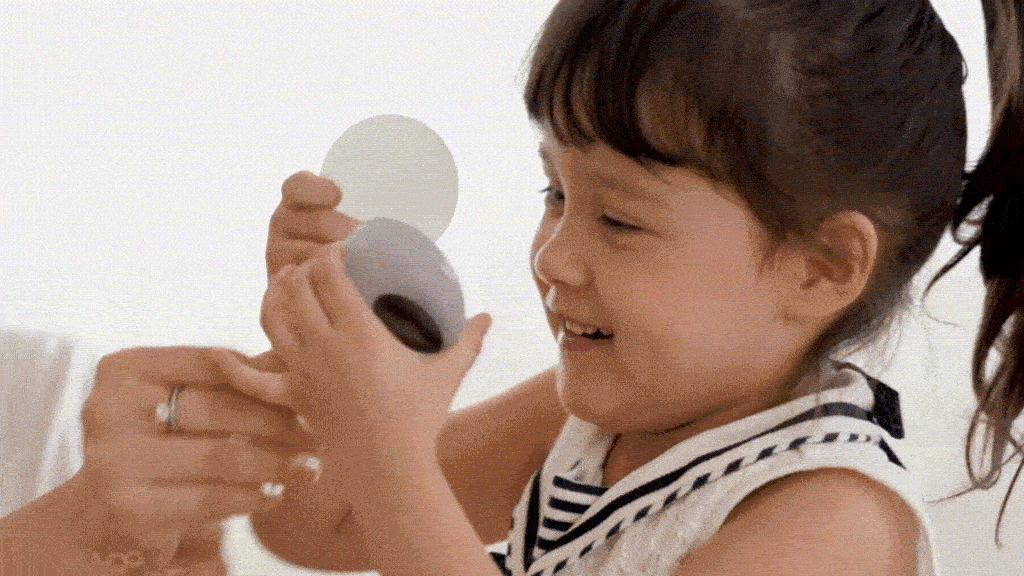 A heartwarming moment unfolds as a young girl, her hair swept up into a high ponytail, beams with excitement. Her white and navy top adds a crisp, cheerful touch to her appearance. The focus of her joy is evident: a small, round box being presented to her. On the left, an adult's hands extend the gift, creating a tender image of generosity. The girl's own hands reach out eagerly, her fingers just about to grasp the box. Her radiant smile captures the pure, unfiltered delight of receiving an unexpected present, embodying the magic of surprise and the simple pleasures of childhood.