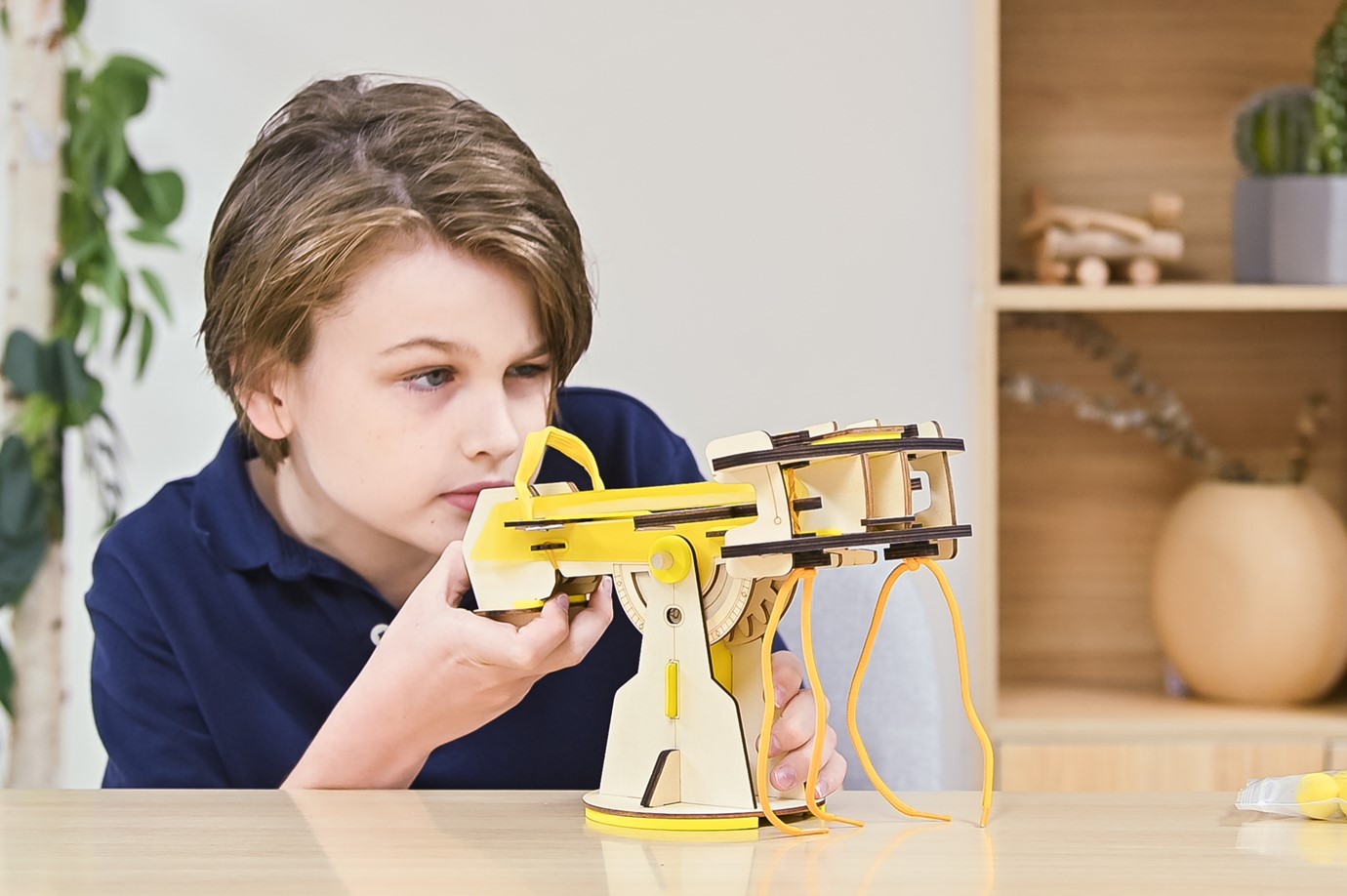 Seated at a table, a young boy with tousled light brown hair leans forward intently, his gaze fixed on a fascinating wooden contraption before him. His navy blue shirt contrasts with the warm tones of the wooden toy, which bears the hallmarks of a carefully assembled construction kit. The device, with its moving parts and bright yellow accents, resembles a miniature crane or catapult. The boy's focused expression and attentive posture reveal his deep engagement with this hands-on exploration of mechanics and design.