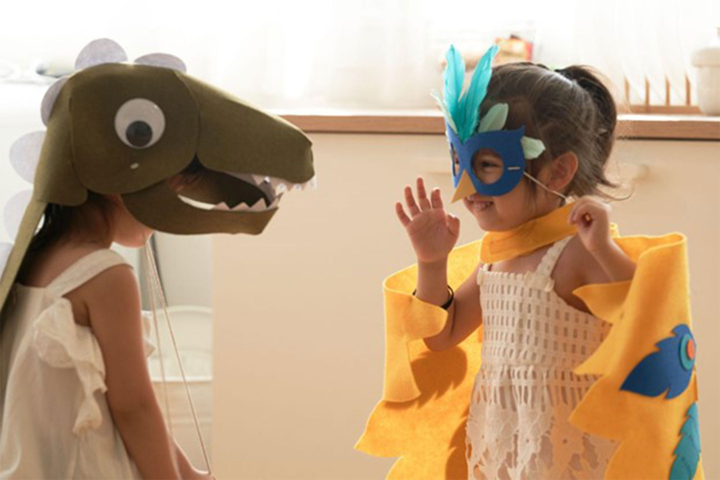 Two young children are engaged in imaginative play. One child wears a large, friendly-looking dinosaur head mask made of felt or similar material and a white sleeveless top. They face the other child, who wears a bright blue bird mask with teal and light green feathers and a yellow cape. The second child also wears a white lacy sundress and has their right hand raised in a gesture. The background is blurred, suggesting they are indoors in a play area or home.
