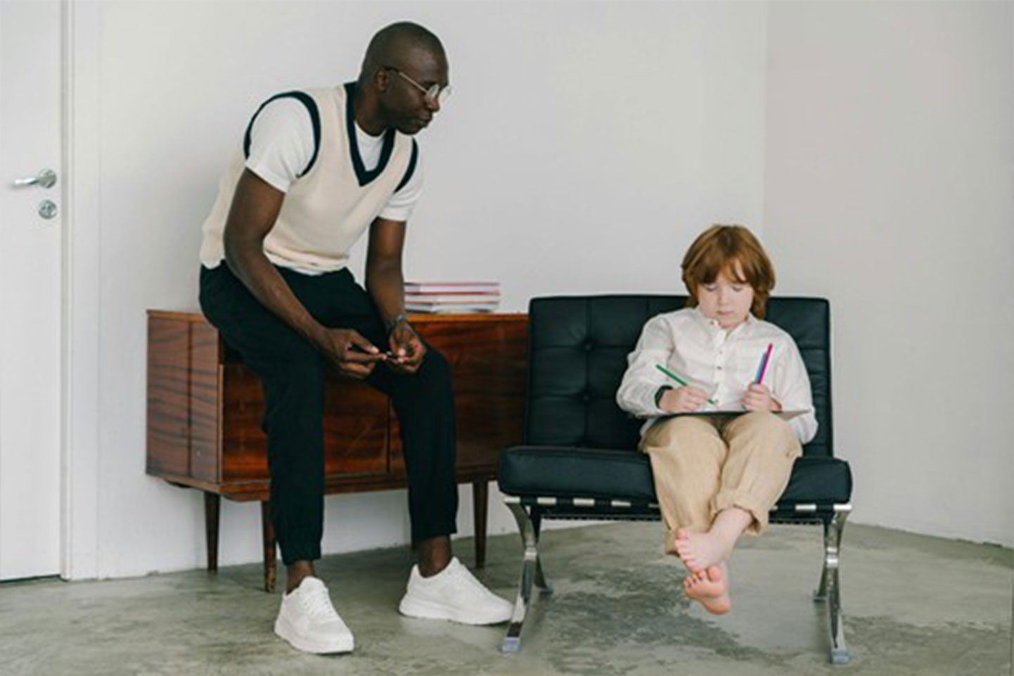 A Black man wearing a cream-colored vest, a white t-shirt, dark pants, and white sneakers sits on a mid-century modern wooden cabinet. He wears glasses and looks towards a child sitting in a black leather and chrome chair. The child, who has reddish hair and wears a white long-sleeved shirt and light brown pants, is barefoot and focused on drawing or writing on a pad of paper, holding colored pencils. The scene appears to be indoors in a minimalist room with white walls and a gray concrete floor. A white door is visible on the left side of the image.