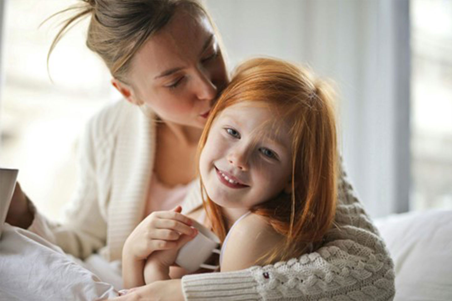 A woman with her hair in a loose bun leans in to kiss a young girl on the head. The woman wears a cream-colored, cable-knit sweater. The girl, who has long, straight red hair, smiles at the camera. She holds a small white cup in her hands. They appear to be sitting on a bed with white sheets. The background is out of focus and appears to be a brightly lit room.