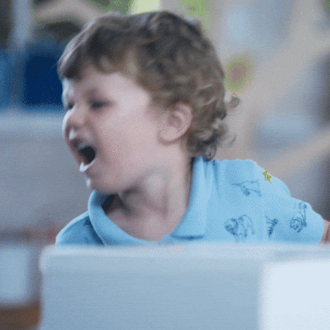 An animated GIF shows a toddler with short, curly brown hair and a light blue collared shirt with animal prints. The child's mouth is open, and they appear to be either speaking animatedly, yelling, or possibly singing. The lower portion of the GIF is blurred, possibly indicating a table or other surface in front of the child. The background is also slightly out of focus, suggesting an indoor setting.