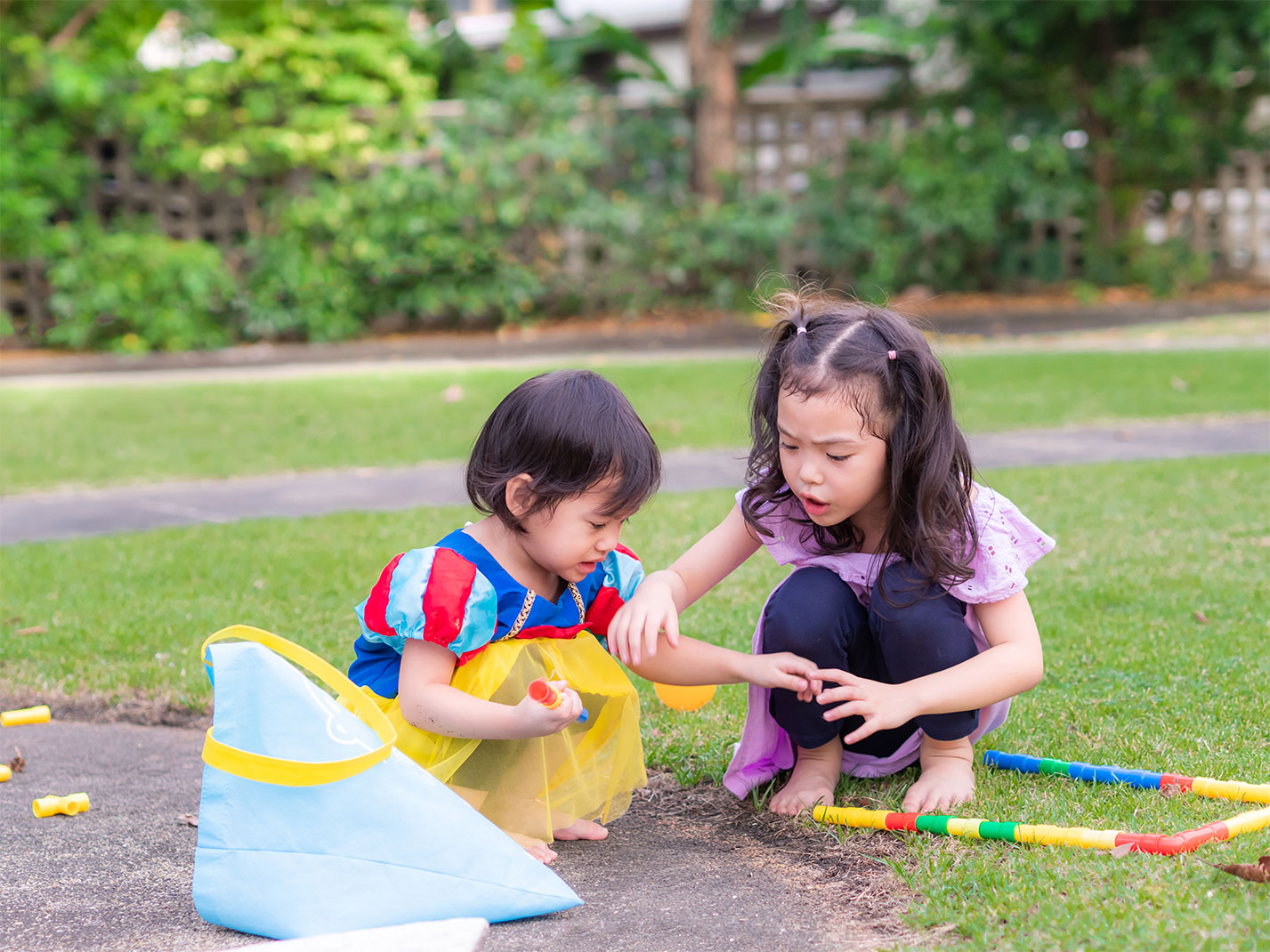 Two young girls are playing outdoors on a grassy area next to a paved path. A toddler, dressed in a Snow White costume with a yellow skirt, sits holding a small toy. An older girl, wearing a light purple top and dark pants, crouches beside her, reaching out and interacting with the younger child. A light blue bag with yellow trim sits near the toddler, and colorful segmented plastic toys are scattered on the grass. The background features green grass, trees, and a glimpse of a fence, indicating a park or backyard setting.