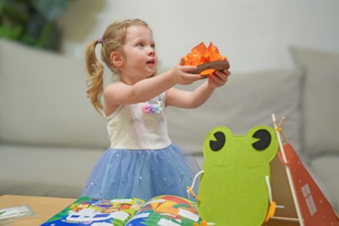A young girl with blonde hair in pigtails, wearing a light-colored sparkly top and a blue tulle skirt, holds a small toy campfire with orange flames above her head. In front of her on a wooden table sits an open book with colorful illustrations, possibly about camping. A stylized felt frog puppet or toy is positioned next to a small toy teepee. A light-colored couch is visible in the softly blurred background.