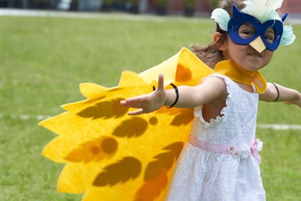 A young girl, wearing a white dress and a bright yellow cape shaped like bird wings, extends her arms as if in flight. She also wears a blue owl mask, complete with white feather accents, which covers her eyes and nose. She appears to be outdoors on a grassy field, likely a park or playground, and smiles at the camera. The cape's design includes darker yellow feather shapes, adding detail to the costume.