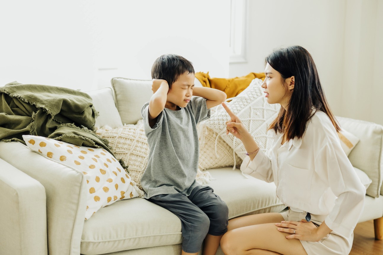 A young boy, wearing a gray shirt and dark pants, sits on a light-colored couch with various pillows and a green blanket. He has his hands over his ears, his face scrunched up in distress. A woman, presumably his mother, kneels beside the couch, wearing a white long-sleeved shirt and light-colored shorts. She points a finger at him, her expression serious and seemingly reprimanding. The scene appears to depict a conflict or disagreement between the mother and child within a living room setting.