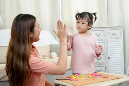 A young child with two small pigtails, wearing a light pink long-sleeved shirt, gives a high five to a woman with long brown hair wearing a peach-colored shirt. The child appears focused on a table in front of them, which holds a brown mat covered with colorful pieces of playdough or modeling clay and a small tool. The woman smiles at the child, engaging in a positive interaction, likely after completing a playdough activity. A light-colored couch and a white cabinet are visible in the softly lit background, suggesting a home or playroom setting.
