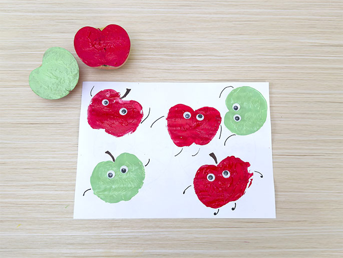 On a light woodgrain table, a white sheet of paper features several red and green apple prints, each adorned with googly eyes and simple, drawn-on arms and legs, giving them the appearance of dancing or moving apples. Above the paper lie two cut apple halves, one red and one green, the tools used to create the prints. This charming scene showcases a completed apple printing art project for children.