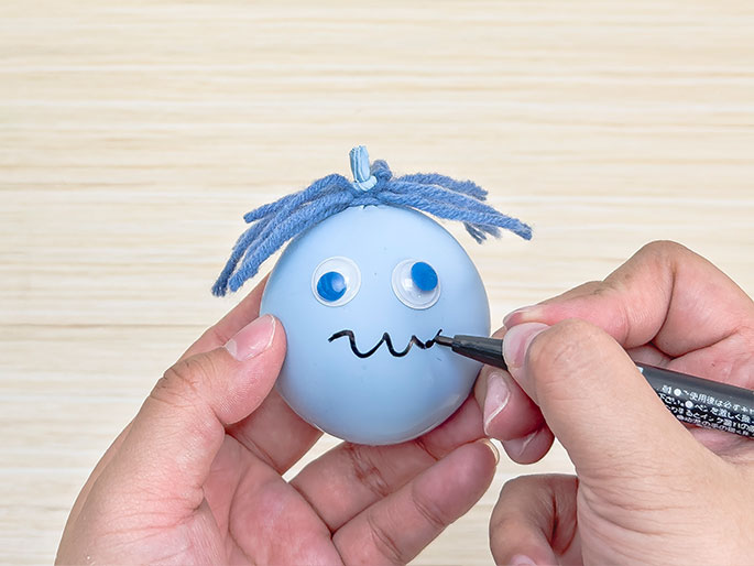 Against a light wood-grain background, two hands are shown completing a balloon creature. The pale blue balloon has a tuft of blue yarn for hair and two googly eyes with bright blue irises. A hand holding a black marker is drawing a wavy, slightly downturned line on the balloon below the eyes to create a whimsical, almost sad-looking mouth. The other hand holds the balloon steady.
