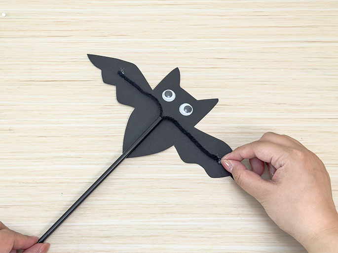 A partially completed bat puppet is shown against a light wood-grain background. The bat, made of black construction paper with googly eyes, has a black stick or straw attached as a handle. A piece of black yarn is strung from the stick and is being held by a hand, which appears to be in the process of attaching it to the bat's wingtips to create a mechanism for moving them.