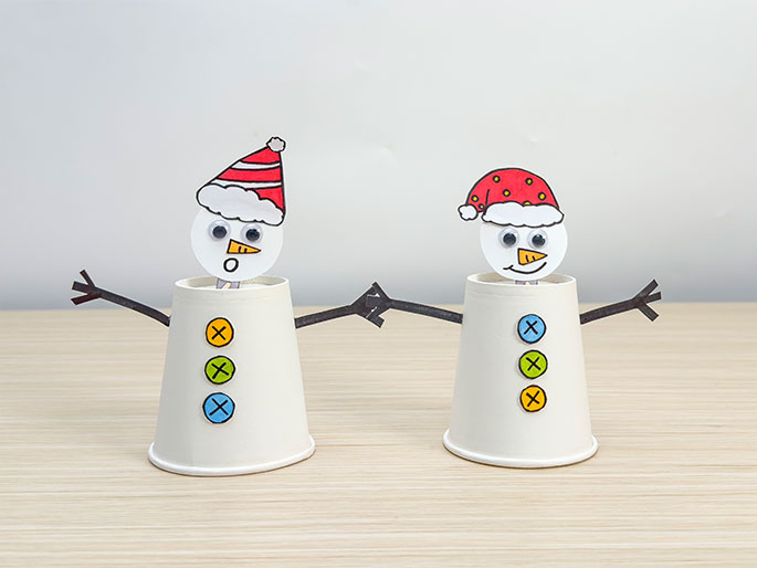 Two adorable snowman figures, crafted from paper cups and paper cutouts, stand on a light wood-grained surface. Each snowman's body is a white cup adorned with three colorful buttons marked with Xs. They have round white heads with googly eyes, carrot noses, and Santa hats. The snowman on the left wears a red and white striped hat and has a surprised expression. The snowman on the right wears a red hat with yellow dots and is smiling. Dark paper arms extend from each snowman, and they appear to be holding hands, completing this festive craft project.