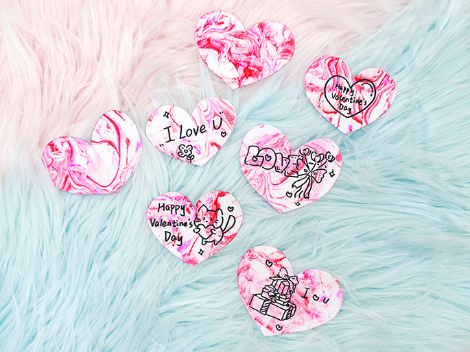 Several hand-decorated Valentine's Day heart shapes are arranged on a fluffy, two-toned background of light pink and teal. Each heart is decorated with a marbled pink and white pattern and features different Valentine's themed messages and drawings, including I Love U, Happy Valentines Day, a cartoon cat, a bow, and presents. The image evokes a sense of handmade crafts and Valentine's Day affection.