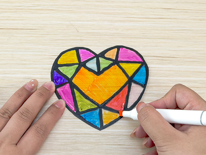 On a light wood-grain surface, two hands are shown coloring a heart-shaped, faceted gem craft. The gem, outlined with black glue or paint, is being filled in with vibrant marker colors within each facet. The colors used include purple, pink, orange, green, light blue, and red, creating a stained-glass effect. One hand holds the gem while the other applies red marker to a lower facet.