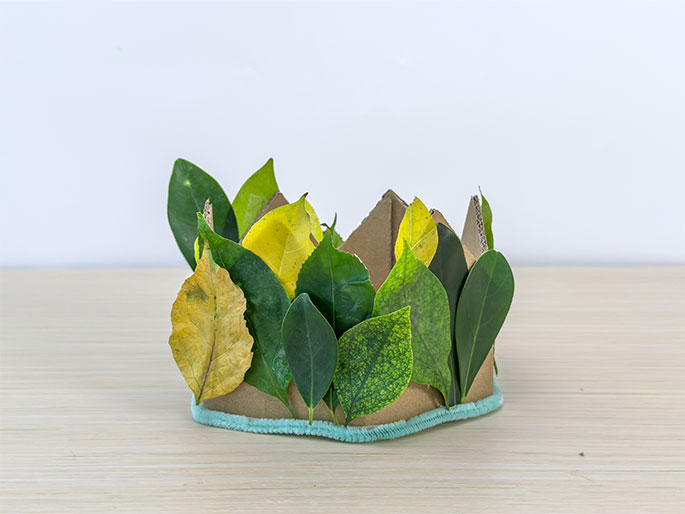 A nature-inspired crown crafted from cardboard and adorned with real leaves rests on a light wooden surface against a plain white background. The crown is made of folded brown cardboard, and a variety of green and yellow-brown leaves are attached to it, giving it a natural, whimsical appearance. A light blue fuzzy or textured trim lines the bottom edge of the crown.