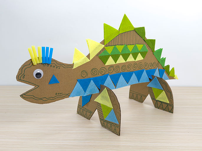 A side view of a playful, handcrafted dinosaur made from cardboard stands on a light wood-grain surface. The dinosaur is primarily brown cardboard, decorated with colorful triangles of light green, bright green, yellow, and various shades of blue tissue paper or felt. These triangles are arranged along its back, sides, and legs. The dinosaur has a row of bright green triangular plates along its spine and a whimsical crest of short, vertical yellow and blue pieces, possibly straws or pipe cleaners, on its head. A googly eye adds to its character. Simple, hand-drawn swirls and lines decorate the cardboard base. The dinosaur's mouth is open, giving it a friendly, almost cartoonish appearance.