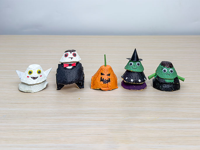Five small Halloween-themed figures crafted from repurposed egg cartons sit on a light wood-grain surface. From left to right, they depict: a friendly white ghost with googly eyes and rosy cheeks; a Dracula figure with a black cape, red bow tie, and fangs; a jack-o'-lantern with a green stem; a witch wearing a black hat and green dress; and a Frankenstein's monster figure in green with black hair. These figures are likely a children's craft project for Halloween decoration.