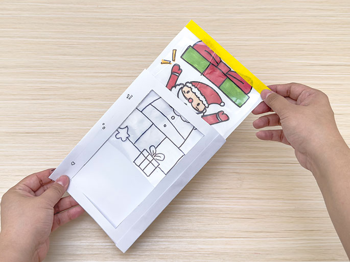 Two hands hold a white card with a cut-out window, partially revealing a coloured drawing of Santa Claus popping out of a present, which is sealed in a plastic bag with yellow tape, against a light wood-grain background.