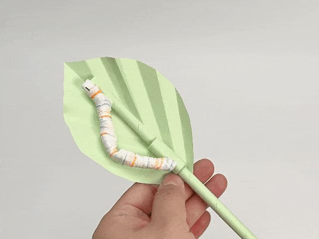 A hand holds a completed craft project featuring a pale green origami leaf attached to a pale green stem. A small, striped paper caterpillar, made with orange, blue, and white stripes, rests on the leaf. The background is a solid light gray or white. This image showcases a finished paper craft, likely depicting a caterpillar on a leaf.