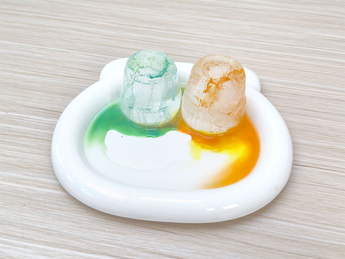 Two small, clear containers, resembling shot glasses, sit on a white, bear-shaped dish placed on a light wood-grain surface. The container on the left holds a vibrant green liquid, which has slightly spilled onto the dish, creating a green puddle. The container on the right holds a bright orange liquid, similarly spilling onto the dish and forming an orange puddle. The containers appear to have some texture or cracking on their surfaces. This image likely depicts a science experiment or a colorful art project involving liquid dyes.