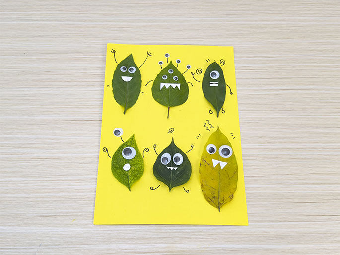 Whimsical leaf monsters crafted on yellow paper. Googly eyes and drawn details bring them to life. A hand adds finishing touches with a marker.
