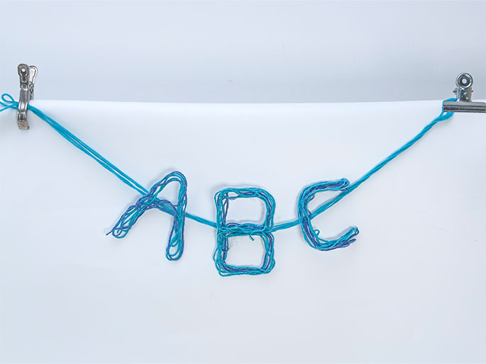 The letters ABC are formed from wrapped teal yarn and strung along a single strand of the same yarn. The yarn is suspended against a white background and held taut by metal clips at each end. The letters have a slightly fuzzy texture due to the nature of the yarn. This appears to be a simple craft project or decorative element.