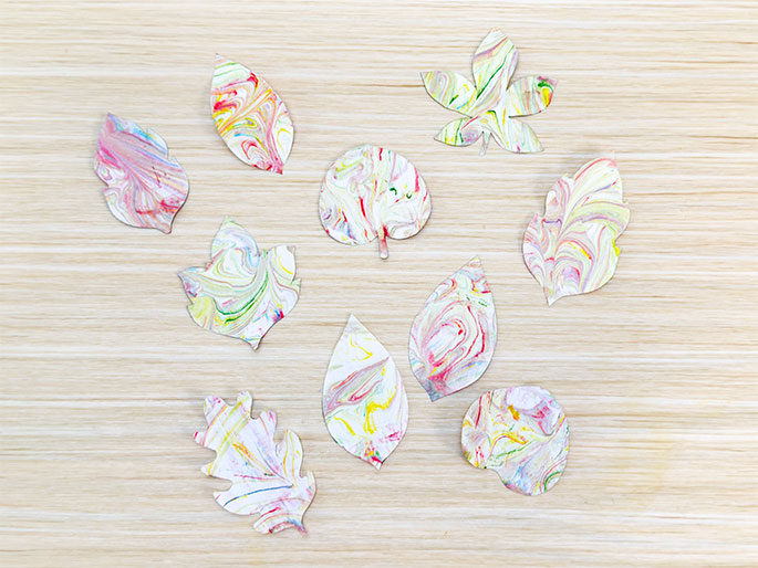 An assortment of paper leaf cutouts, each decorated with a marbled pattern of swirling pink, yellow, blue, and green paints, lies on a light woodgrain surface. The leaves vary in shape and size, resembling different types of leaves. This image likely depicts the result of a marbling art project.