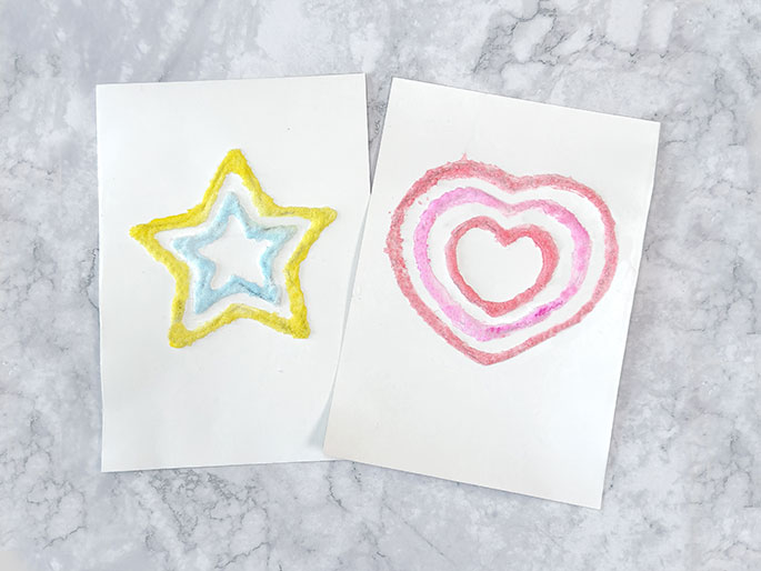Two pieces of white paper sit on a marbled surface. The paper on the left has a star shape made from layers of colored salt or sand, with yellow on the outside, then blue, and white in the center. The paper on the right has a heart shape made with similar layered colors, using shades of pink and a white center. The image showcases a simple art activity using colored granular materials.