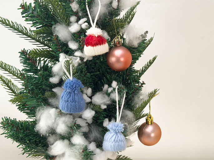 A small, artificial Christmas tree is decorated with miniature knitted hats and shiny rose-gold ornaments. The hats are red and blue, each topped with a pom-pom. Bits of white fluffy material, likely representing snow, are tucked among the branches. The tree is set against a plain, off-white background.