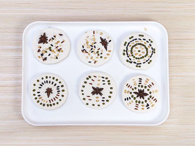 Six round, flat objects, possibly made of dough or clay, are arranged on a white rectangular tray. Each object is decorated with a variety of small seeds and spices, creating different patterns. Some feature star anise at the center, while others have concentric circles or radiating lines of various colored seeds. The tray rests on a light wood-grained surface. This image likely depicts a craft or culinary activity.