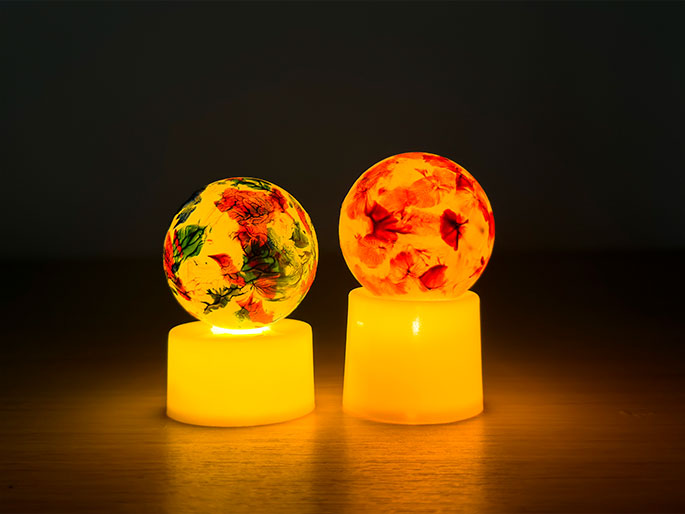 Two decorative illuminated spheres sit on a wooden surface against a dark background. Each sphere rests on a short, cylindrical base that emits a warm, yellow light. The spheres themselves are translucent and contain embedded floral patterns. The sphere on the left has a mix of green, orange, and red floral designs, while the sphere on the right features primarily orange and red floral patterns. The light from the bases illuminates the spheres from below, highlighting the floral designs and creating a soft, ambient glow.