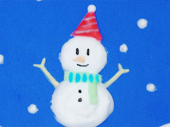 A cheerful snowman crafted from a white, fluffy material is depicted against a bright blue background. The snowman wears a red and white striped conical hat and a light green scarf with blue dashed lines, suggesting a knitted pattern. Small, white dots scattered around the snowman represent falling snow. The snowman has simple facial features drawn on, including eyes, a carrot nose, and a smiling mouth, and small, light-colored stick arms raised in a welcoming gesture. This image likely represents a winter-themed craft project.