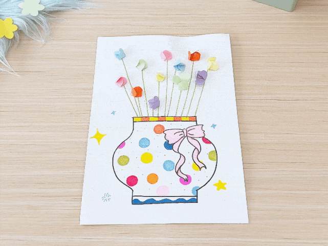 A crafting video showcases the creation of a colorful flower arrangement in a vase. The vase, drawn on white paper, is decorated with colorful polka dots and a pink ribbon. Small, pastel-colored paper flowers are attached to green stems, creating a bouquet that sits within the drawn vase. The background is a plain wooden surface. The video focuses on the finished artwork, highlighting the vibrant colors and simple design.