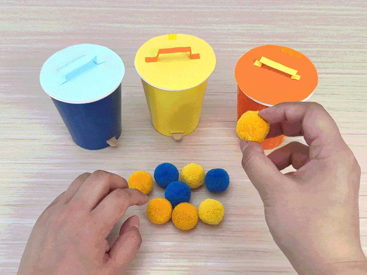 Two hands interact with three small, colorful bins on a light wooden table. The bins, resembling miniature trash cans, are made from paper cups with light blue, yellow, and orange lids and attached handles. Several small, fluffy pom-poms in blue and yellow are clustered near the bins. The hands pick up yellow pom-poms one at a time and place them into the corresponding yellow bin, demonstrating a color-matching or sorting activity.