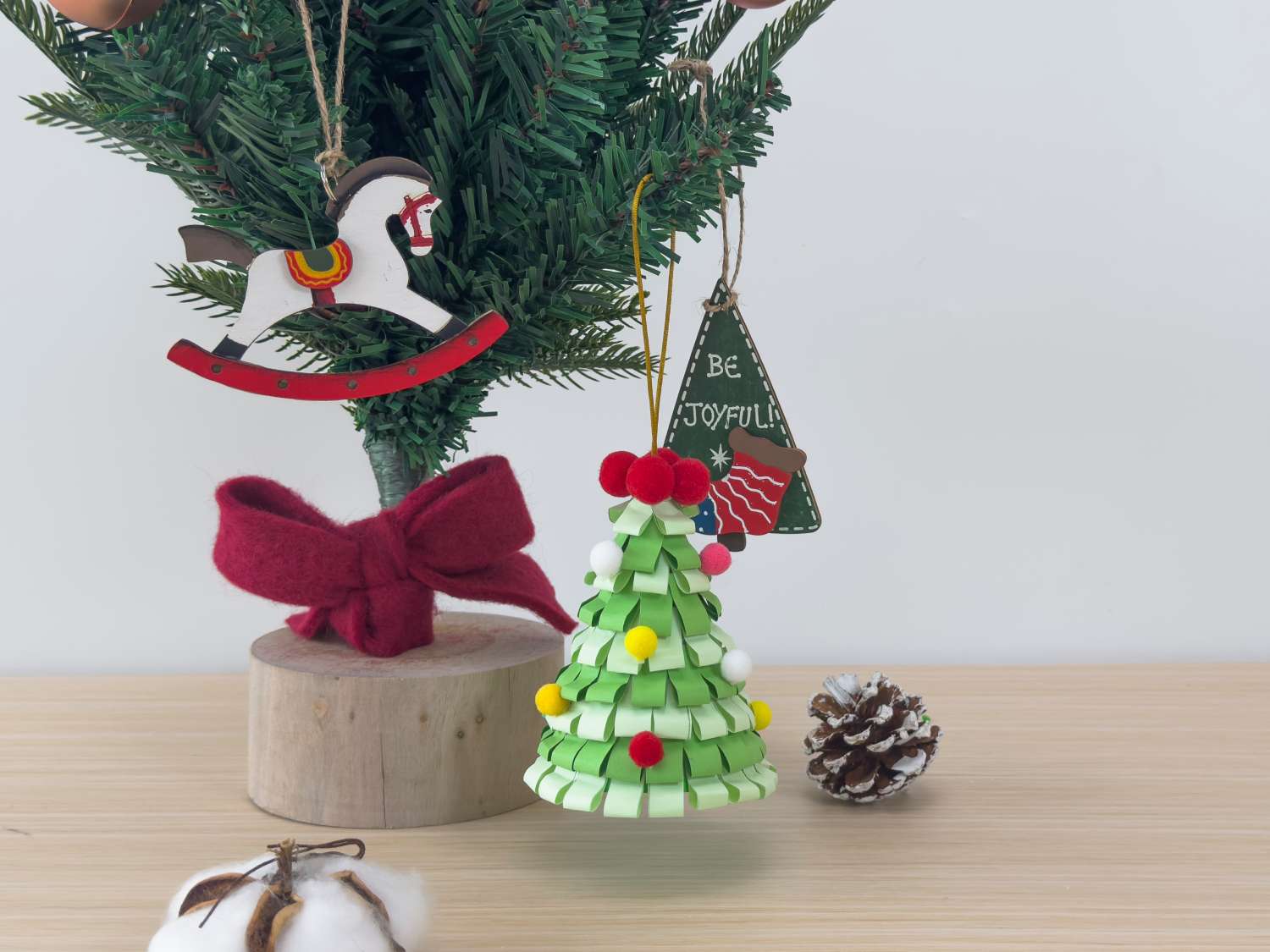A small artificial Christmas tree decorated with handmade paper ornaments, a wooden rocking horse, and a Be Joyful sign sits on a wooden surface alongside a pine cone and cotton boll.