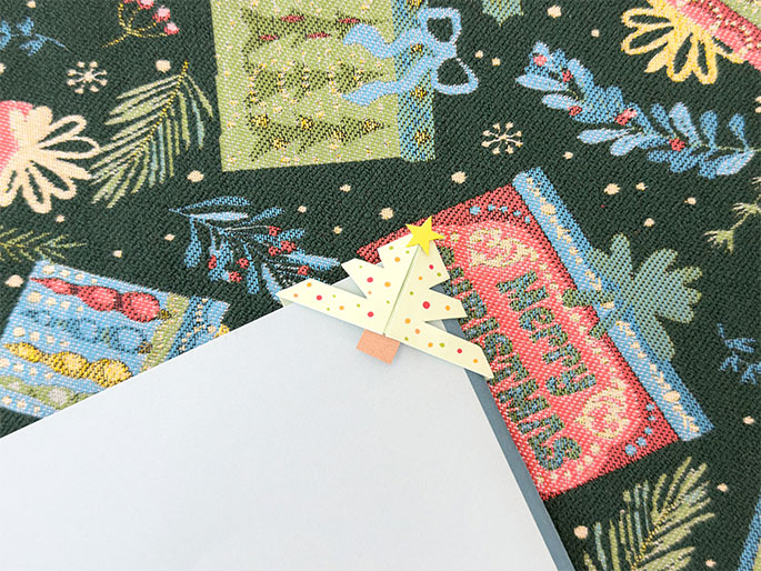 A light blue piece of paper or card stock rests on a dark green fabric printed with Christmas motifs like presents, trees, and snowflakes. Attached to the corner of the blue paper is a small, folded paper Christmas tree. The tree is pale yellow-green with small red and yellow dots and a small gold star at the top. A small rectangular piece of orange paper acts as the tree's trunk. The image shows a festive, handmade Christmas tree corner bookmark.