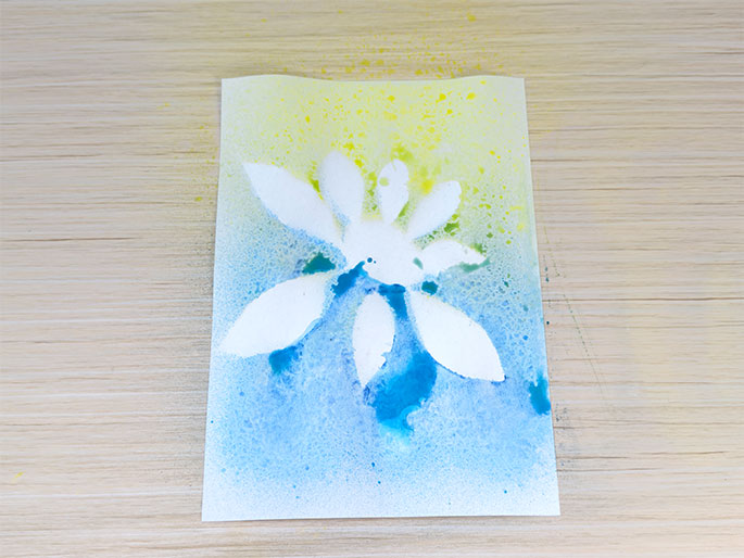 A piece of white paper, resting on a light wood-grain surface, displays a flower-like silhouette in white against a backdrop of sprayed blue and yellow paint. The top portion of the paper is predominantly yellow, while the bottom is primarily blue, with some colour overlap in the middle. The white flower shape, created by masking with leaves and a rock, stands out against the colourful, speckled background.
