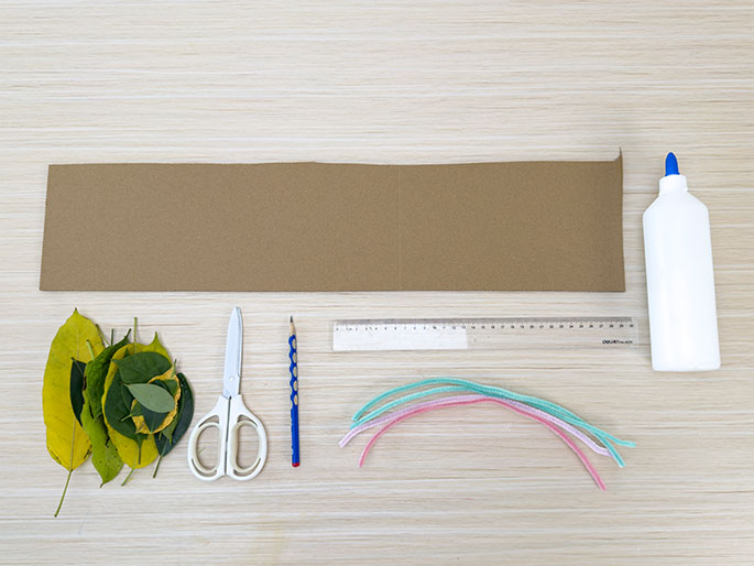 An overhead flatlay image displays the materials needed for a craft project arranged on a light woodgrain surface. The materials include a rectangular piece of brown cardboard, a small pile of green and yellow leaves, a pair of white-handled scissors, a pencil, a clear plastic ruler, several colorful pipe cleaners in pink, light green, and light blue, and a white bottle of glue.