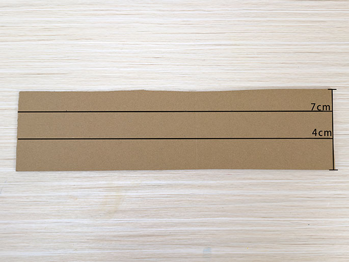 A rectangular piece of brown cardboard lies flat on a light woodgrain surface. Two horizontal lines are drawn across the cardboard, dividing it into three sections. Labels indicate the dimensions of the sections: 7cm for the top section and 4cm for the middle section. The bottom section's dimension is not labeled, but it appears to be the same size as the middle section (4cm). This image likely illustrates the first steps in preparing the cardboard for a craft project.