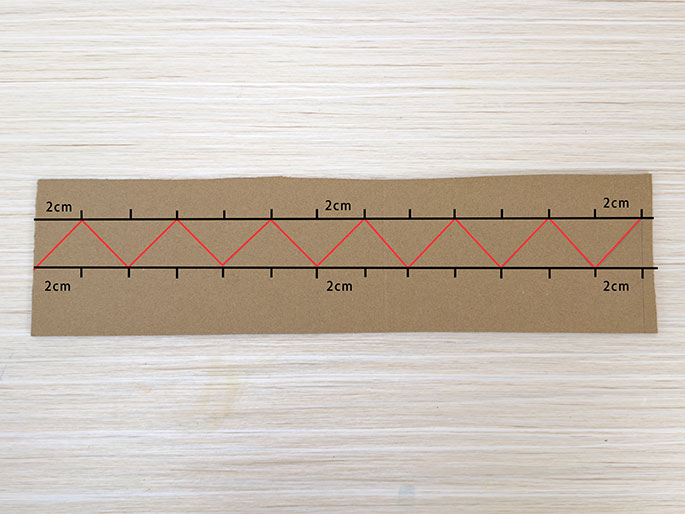 A rectangular piece of brown cardboard rests on a light woodgrain surface. Black lines mark 2cm increments along the top and bottom edges of the cardboard. Connecting these marks are red lines forming a series of connected triangles or a zig-zag pattern across the cardboard. The 2cm labels indicate the length of each segment along the top and bottom edges, as well as the height of each triangle. This image likely demonstrates marking the cardboard for cutting or folding in a craft project.