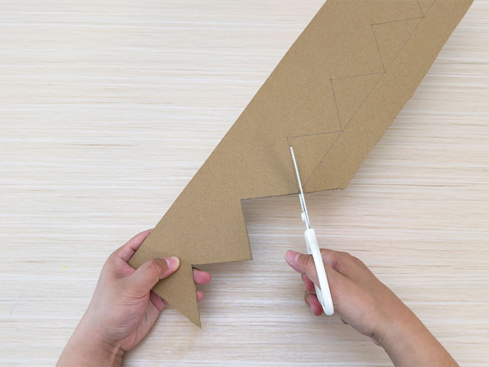 Two hands work on a piece of brown cardboard on a light woodgrain table. One hand holds the cardboard, which has been marked with a zig-zag pattern and partially cut. The other hand holds a pair of white-handled scissors, actively cutting along one of the marked lines. The cutting process is creating a series of triangular points along the edge of the cardboard. The image showcases the process of cutting out a shape from cardboard, likely for a craft project.