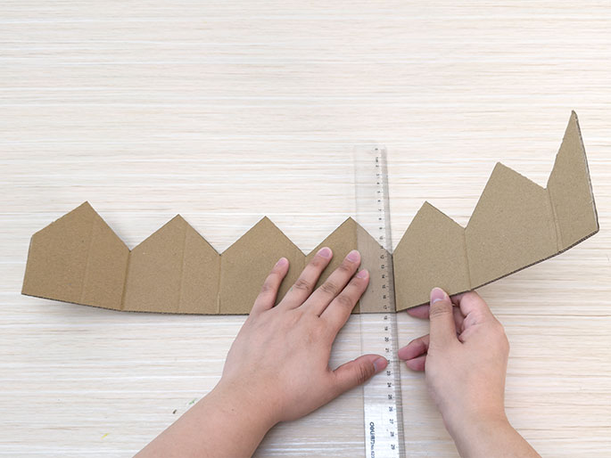 Two hands manipulate a piece of brown cardboard cut into a crown shape with triangular points along the top edge on a light woodgrain surface. The hands are using a clear plastic ruler to make a fold along one of the scored lines between the triangular points. The image demonstrates the process of shaping the cardboard crown.