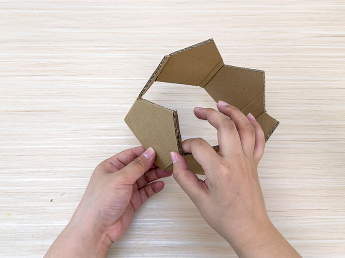 Two hands hold a piece of brown cardboard that has been folded and shaped into a circular crown form, on a light woodgrain surface. The crown has a jagged, multi-pointed top edge. The hands are in the process of joining the two ends of the cardboard together, preparing to overlap and glue them to complete the crown. The image captures the stage of assembling the cardboard crown where the final connection is being made.