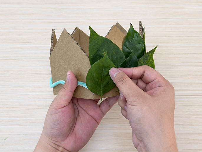 Two hands hold a simple crown made of brown cardboard. A light teal fuzzy yarn or pipe cleaner is glued along the bottom edge of the crown. Green leaves are being added to the crown; one hand holds the crown while the other positions the leaves. The image shows the final stages of creating a nature-inspired crown, likely for a craft or play activity.