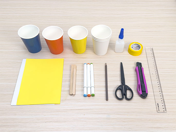 An assortment of craft supplies lies arranged on a light woodgrain table. Four paper cups in blue, orange, yellow, and white are positioned at the top left. A bottle of white glue and a roll of yellow tape are next to the cups. A piece of bright yellow construction paper sits below the blue cup. Three white markers with orange, green, and blue lids are grouped together, alongside two wooden craft sticks and a brown pencil. Black-handled scissors, a magenta utility knife, and a clear plastic ruler complete the collection of supplies. The image displays the materials gathered for a craft project.