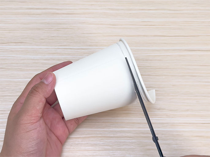 A hand holds a white paper cup against a light woodgrain background. Scissors are cutting along the rim of the cup, removing a thin strip of the top edge. The image shows the process of trimming or modifying a paper cup using scissors, likely as part of a craft or DIY project.