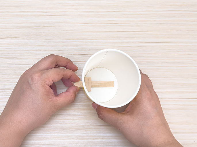 Two hands hold a white paper cup over a light woodgrain background. Inside the cup, a small wooden craft stick is positioned horizontally. One hand holds the cup while the other adjusts the craft stick. The image shows the process of assembling a craft project using a paper cup and a wooden stick. The cup appears to be prepared for some sort of insertion or attachment related to the stick.
