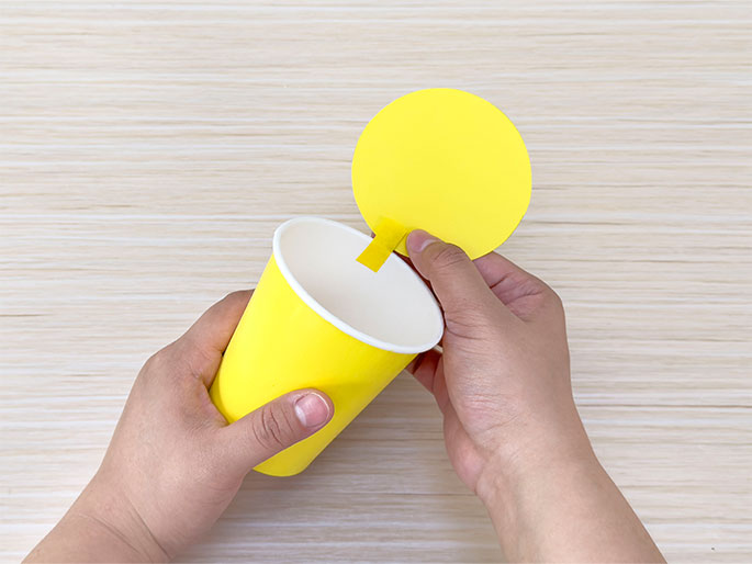 Two hands hold a bright yellow paper cup and a yellow circle of paper over a light woodgrain background. The circle has a small piece of tape attached to it and is being inserted into the cup. The image shows the process of creating a craft project using a paper cup and a paper circle, likely a lid or a decorative element being added to the cup.