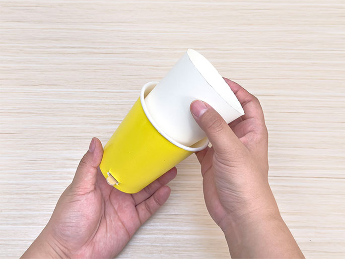 Two hands hold a bright yellow paper cup with a rectangular slot cut into its side and a plain white paper cup against a light woodgrain background. The white cup is positioned slightly inside and on top of the yellow cup, as if being placed over it. The image shows two paper cups being used in a craft project, likely being combined or modified in some way.
