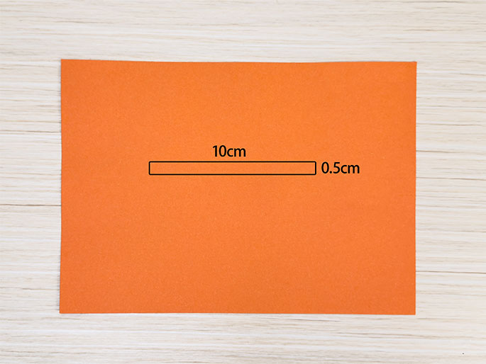 A rectangular piece of bright orange construction paper lies on a light woodgrain background. A narrow, rectangular section is marked near the center of the paper, labeled with the dimensions 10cm in length and 0.5cm in width. The image illustrates a measured and marked section on a piece of paper, likely for cutting or as a guide for a craft or construction project.