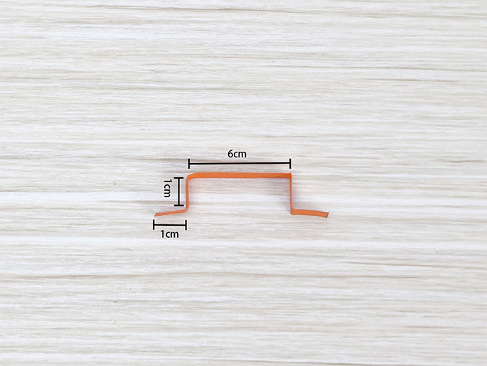 A small, orange strip of paper, bent into a Z or elongated S shape, lies on a light woodgrain background. Lines and labels indicate the dimensions: a 6cm long horizontal section, with two 1cm vertical sections at each end, creating the bends. The image shows a piece of folded or bent paper, likely a component for a craft or paper construction project.